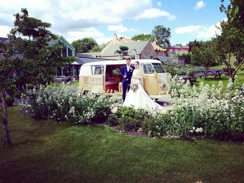 Married couple posing beside camper for photo holding vw campervan champagne flutes