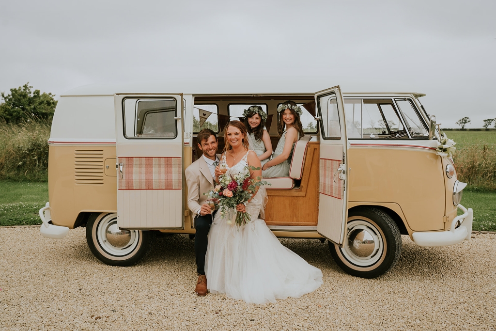 A married couple and two daughters looking out of the side doors of a vw splitscreen campervan posing for a family photo at the wedding